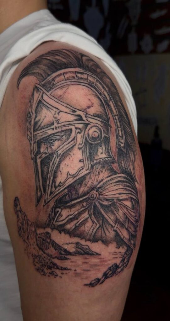 Black & Grey Tattoo am Oberarm: Spartaner-Helm mit Schattierung